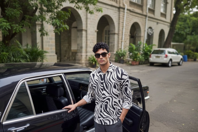 A man wearing a black and white wavy patterned button-up shirt and dark gray trousers stands next to an open black car door on a street.